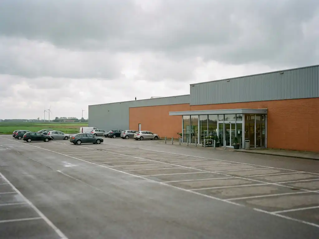 Large-format retail warehouse store with vast parking lot, flat Dutch polder landscape on the horizon under overcast skies.
